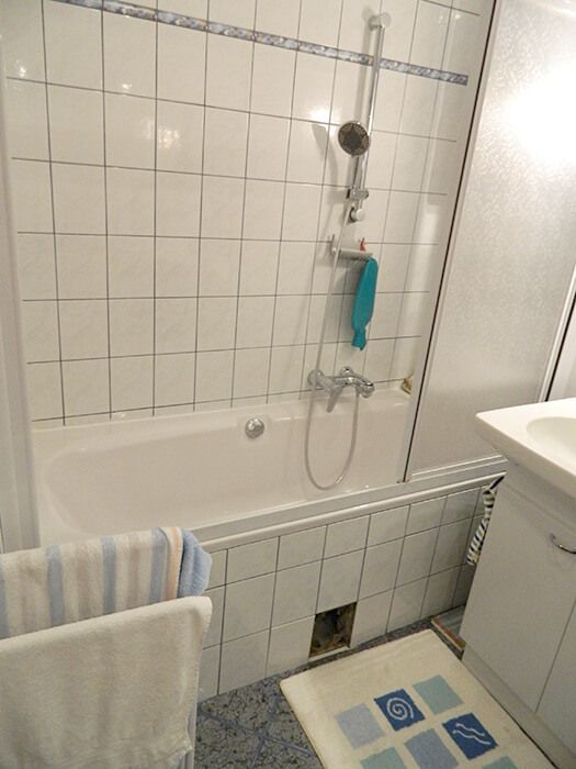 Älteres Badezimmer mit Badewanne und Duschvorrichtung, weißen Wandfliesen und Handtuchhalter.