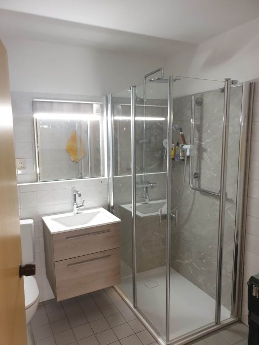 Modernisiertes Badezimmer mit neuer Glasdusche, hellgrauen Wandfliesen in Steinoptik, hängendem Waschtisch mit Holzfronten und beleuchtetem Spiegel. Die cleane, helle Gestaltung wirkt hochwertig und funktional – Referenz einer Teilsanierung durch HSA Scharrenbroich.