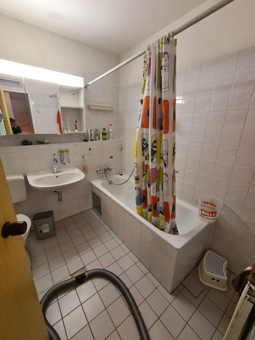 In die Jahre gekommenes Badezimmer mit Badewanne, buntem Duschvorhang, klassischem Waschbecken und sichtbaren Alltagsgegenständen. Der Raum zeigt typische Gebrauchsspuren – Zustand vor der Teilsanierung durch HSA Scharrenbroich.